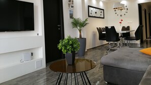 Un salon moderne avec un téléviseur à écran plat, une table ronde en verre, un canapé gris, une porte noire, une plante en pot, un coin repas avec une table blanche et des chaises noires, et une horloge murale.