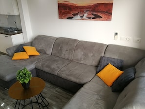 Moderni olohuone, jossa on harmaa sohva, kaksi oranssia koristetyynyä, pieni pyöreä pöytä, jossa on kasvi, ja seinällä oleva maalaus, jossa on kuvattuna kanjoni.