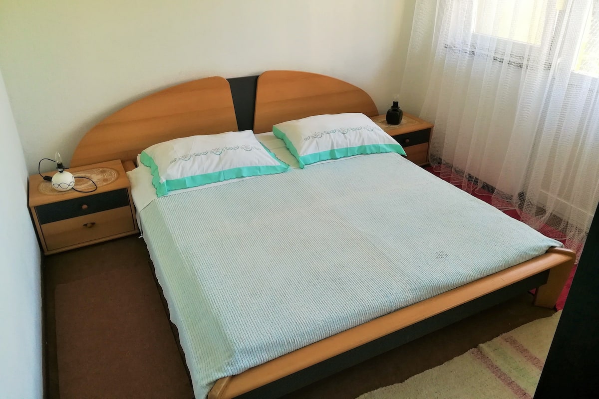Een enkel bed met houten hoofdbord, twee kussens, een nachtkastje met een lamp en een nachttafeltje.