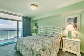 A bedroom with a large bed, a nightstand with a lamp, a framed picture on the wall, and a view of the ocean through a large window.