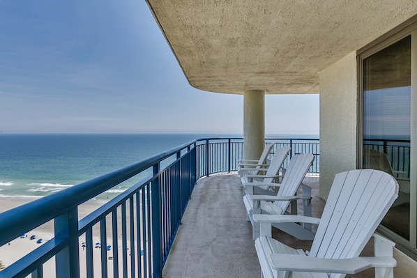 A balcony with a view of the beach and ocean. There are white Adirondack chairs and a blue railing.