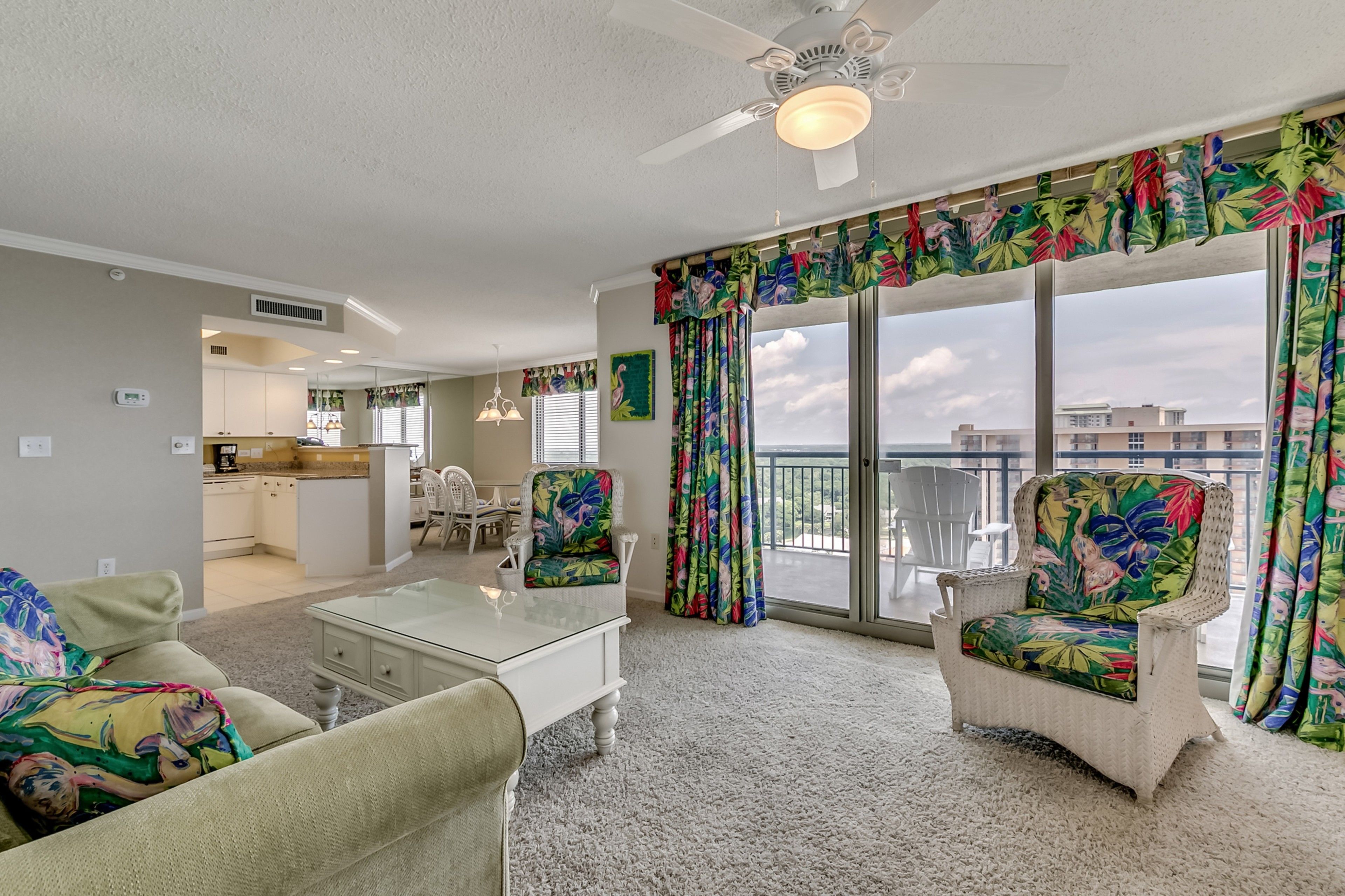 A spacious living room with a glass coffee table, two wicker chairs, a sofa, and a balcony with large windows and tropical curtains.