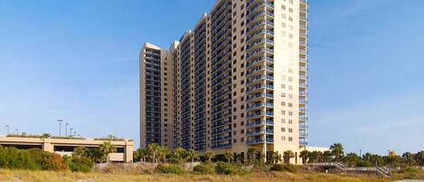 A high-rise hotel with multiple balconies, located near a beach with a boardwalk.