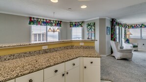 Eine Küche mit Granit Arbeitsplatte, weißen Schränken und dekorativen, sternförmigen Griffen. Es gibt Fenster mit Jalousien und bunten, tropischen Vorhängen.