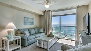 A modern living room with a sofa, a coffee table, a side table with a lamp, a ceiling fan, a TV, and a large window with a view of the ocean.