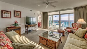 A spacious living room with a large flat-screen TV, comfortable seating, a glass coffee table, and a balcony with a view of the ocean.