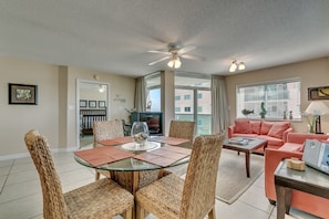 A modern living room with a glass-top dining table, wicker chairs, a ceiling fan, and a pink sofa. - Crescent Keyes by Condo-World (North Myrtle Beach)