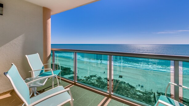 A balcony with two chairs and a glass railing overlooking a beach with clear blue water and a distant shoreline.