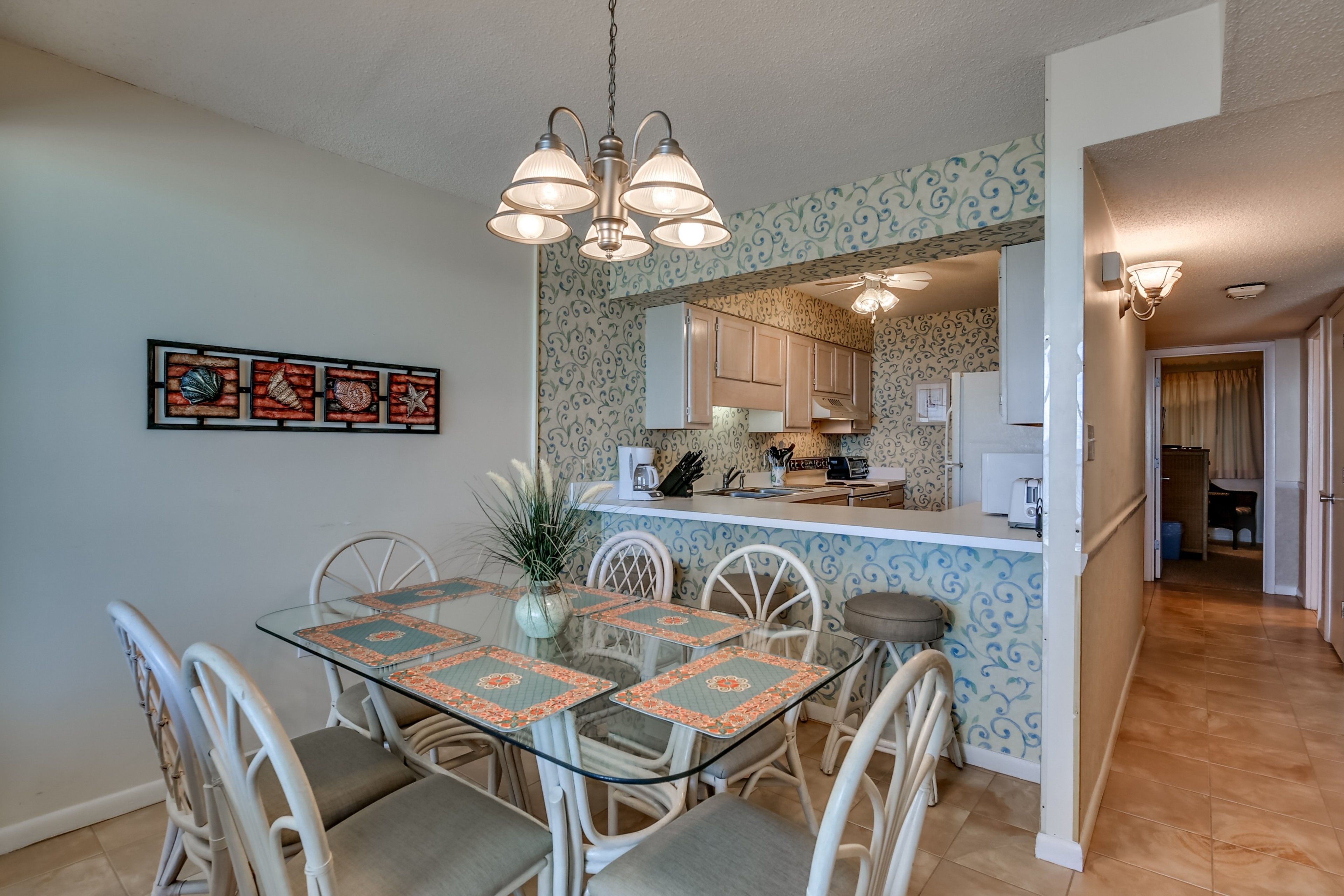A dining area with a glass table and chairs, a hanging light fixture, and a kitchen in the background.