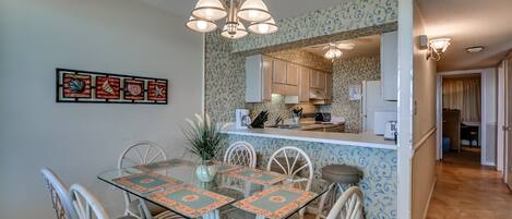 A dining area with a glass table and chairs, a hanging light fixture, and a kitchen in the background.