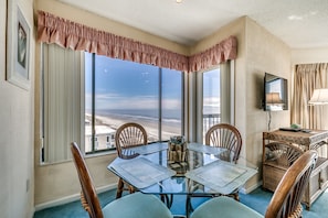 A dining area with a glass table, wooden chairs, and a view of the beach. - Crescent Towers by Condo-World (North Myrtle Beach)