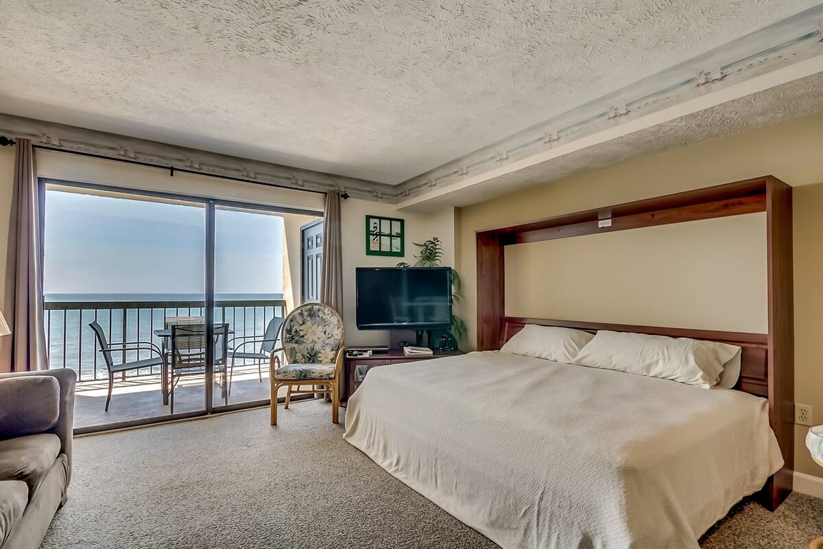 A hotel room with a large bed, a TV, a desk, and a chair. There is a balcony with a view of the ocean.
