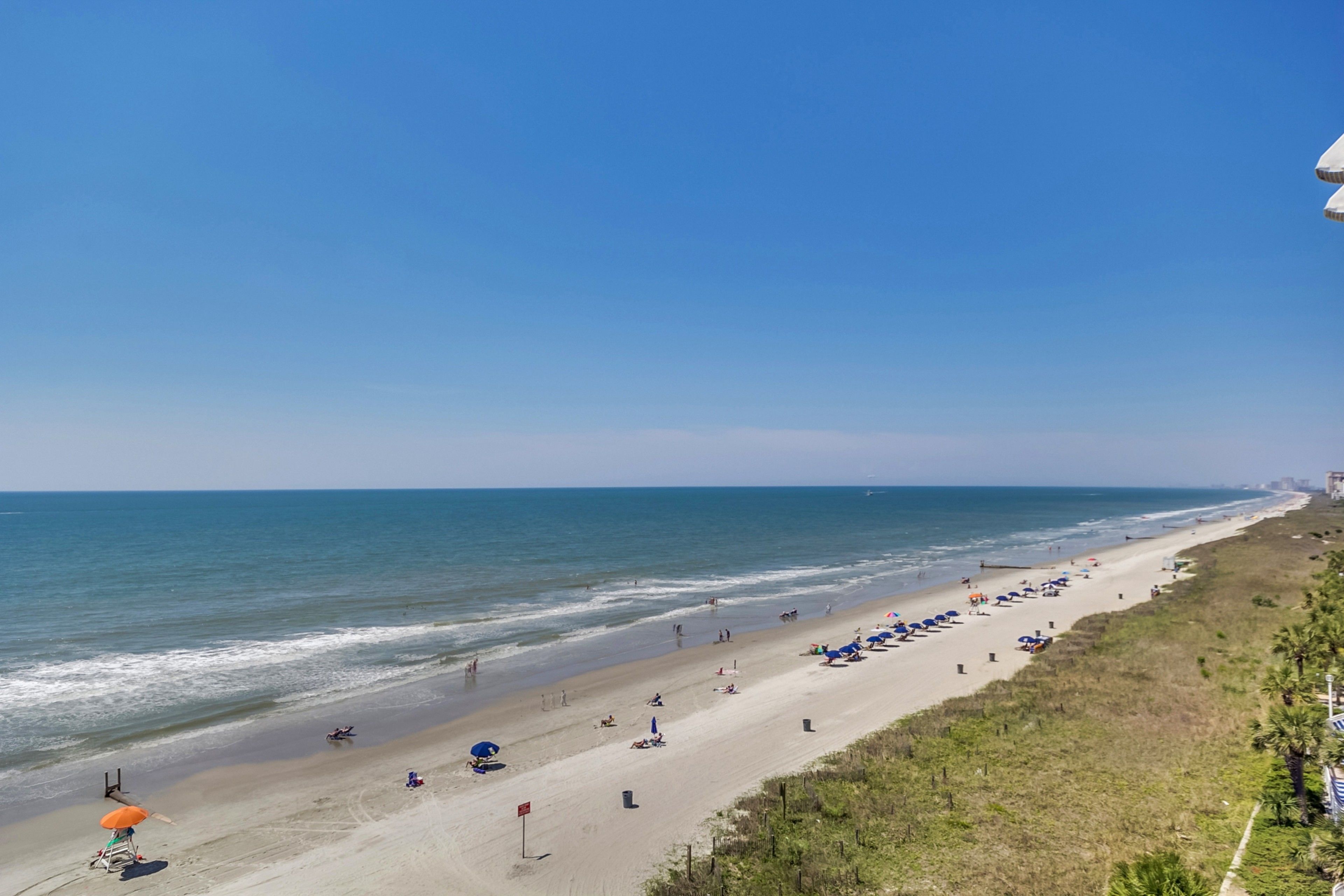 A beach with umbrellas and people, a clear sky, and the ocean.