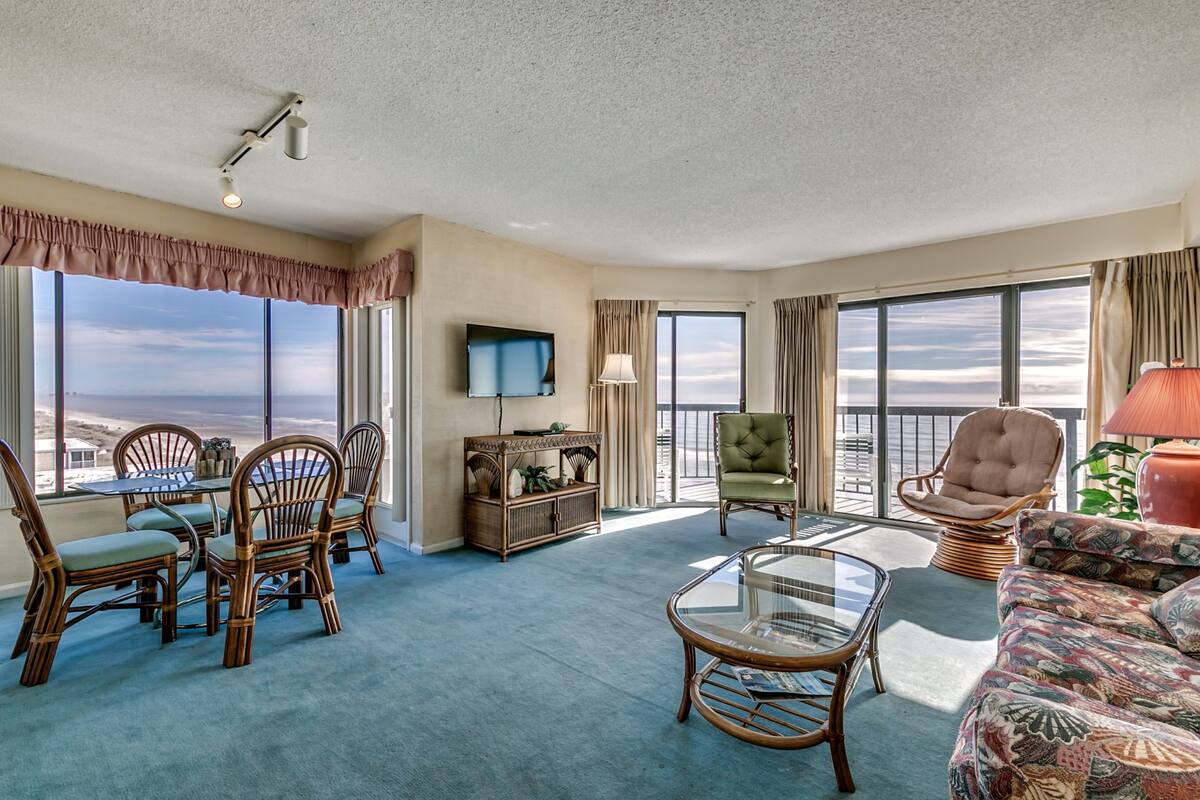 A spacious living area with a dining set, a television, and a balcony with a view.