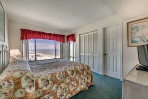 A bedroom with a large bed, a window with a view of the beach, a door, and a television on a stand. - Crescent Towers by Condo-World (North Myrtle Beach)