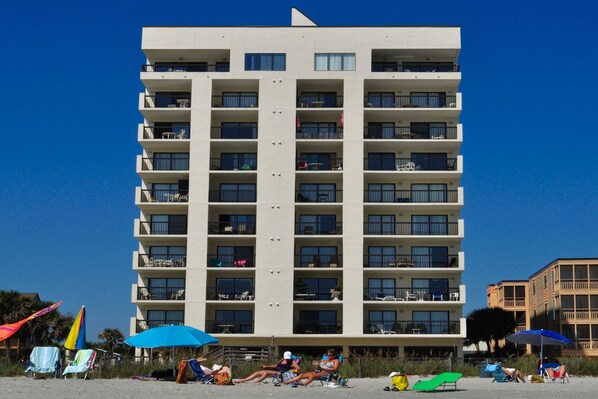 A multi-story hotel with balconies, a beach in the foreground, and a clear blue sky.