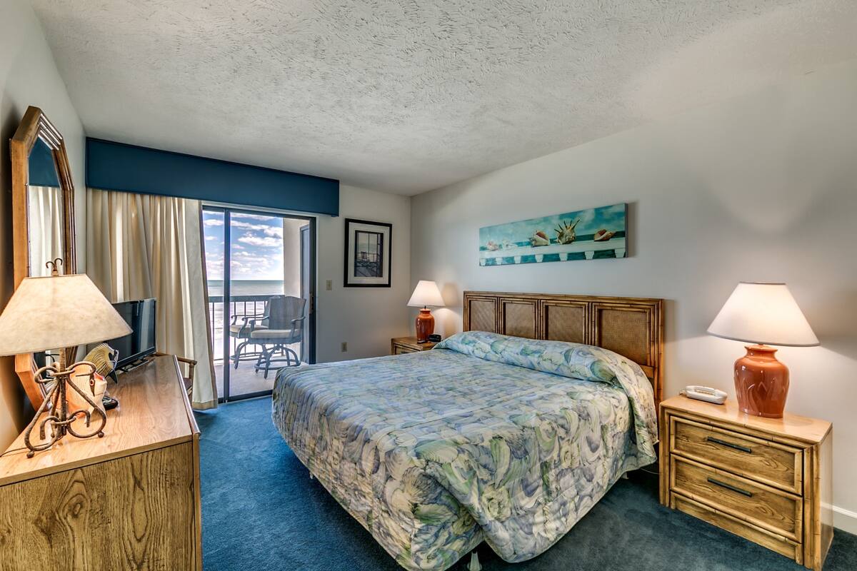 A hotel room with a large bed, two bedside lamps, a wooden nightstand, a desk with a lamp, a chair, and a view of the ocean through a sliding door.