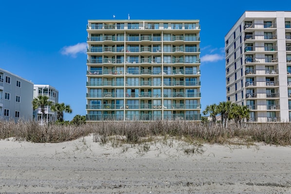 A multi-story hotel with balconies, located near a sandy beach area.