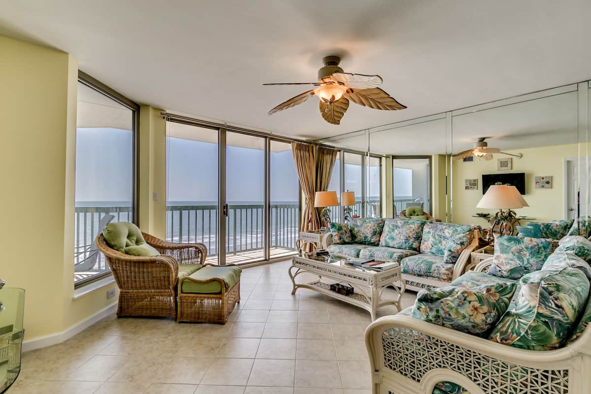 A spacious living room with a balcony, wicker furniture, a patterned sofa, and a ceiling fan.