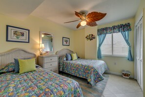 A bedroom with two beds, a ceiling fan, a window with curtains, and a small table with a lamp.