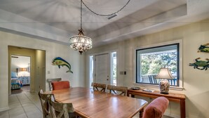 A dining area with a wooden table, chairs, a chandelier, and a window with a view of trees. - High C's by Condo-World (North Myrtle Beach)