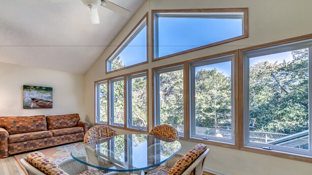 A spacious room with a glass-top dining table surrounded by wicker chairs, a patterned sofa, and large windows offering a view of trees. - High C's by Condo-World (North Myrtle Beach)