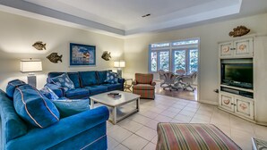 A spacious living room with a blue sofa set, a coffee table, and a striped ottoman. There is a television on a white cabinet, and a window with a view of trees. - High C's by Condo-World (North Myrtle Beach)