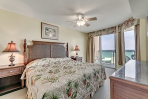 A hotel room with a bed, two nightstands with lamps, a ceiling fan, a framed picture, a balcony with a view of the ocean, and a glass-top table.