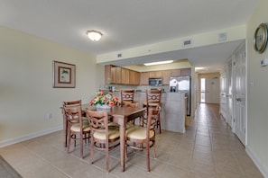 A dining area with a wooden table and chairs, a kitchen in the background, and a hallway leading to a door.