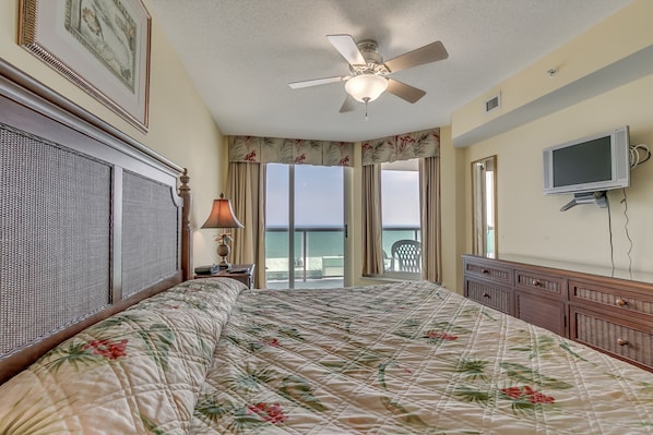 A bedroom with a large bed, a ceiling fan, a TV, and a view of the ocean.
