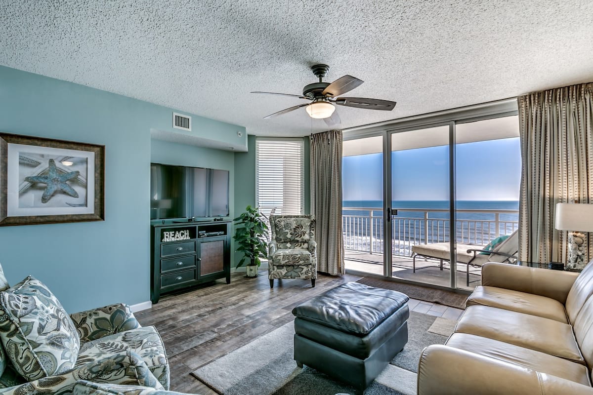 A spacious living room with a large window offering a view of the ocean, a ceiling fan, and a television.