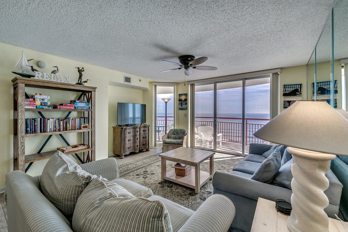 A living room with a sofa, armchair, and coffee table, a bookshelf, and a view through large glass doors.