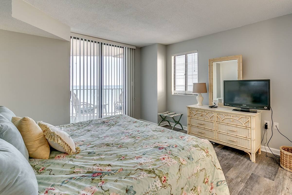 Ein Schlafzimmer mit Bett, Kommode, Fernseher und einem Fenster mit Jalousien.