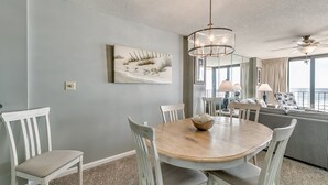 A dining area with a round wooden table, white chairs, and a chandelier. A painting of grass and birds hangs on the wall. - Summit by Condo-World (North Myrtle Beach)