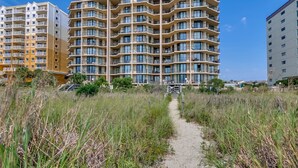 A multi-story residential building with balconies, a sandy path leading to it, and surrounding grassy areas. - Summit by Condo-World (North Myrtle Beach)