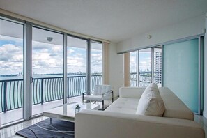 A modern living room with a white sofa, a small white table, and a chair. Large floor-to-ceiling windows offer a view of a city skyline and a body of water. - Spectacular Midtown Miami (Miami)