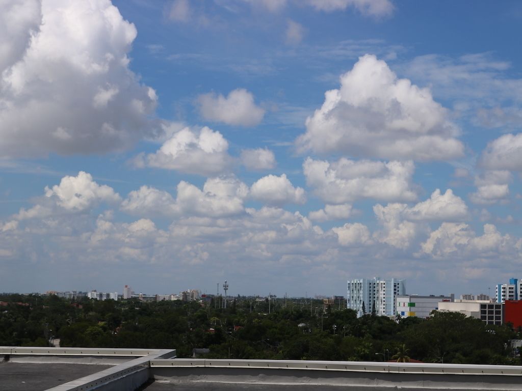 A cityscape with a clear sky and clouds, featuring modern buildings and a green area in the foreground.