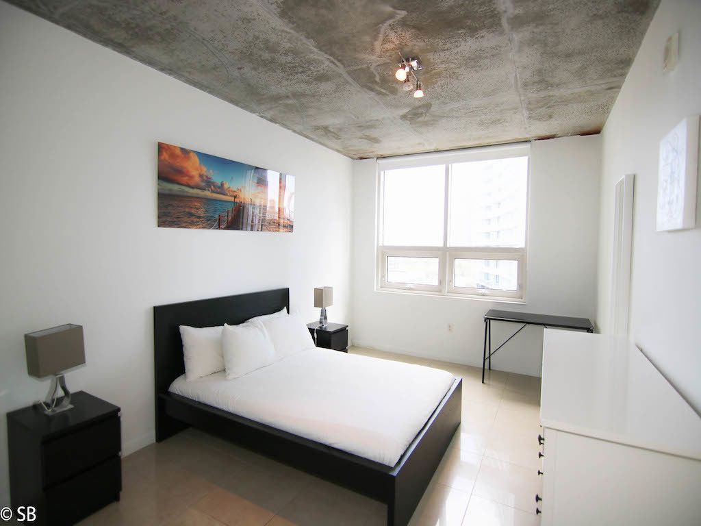 A modern bedroom with a large bed, a nightstand, a desk, and a window with a view of the city.