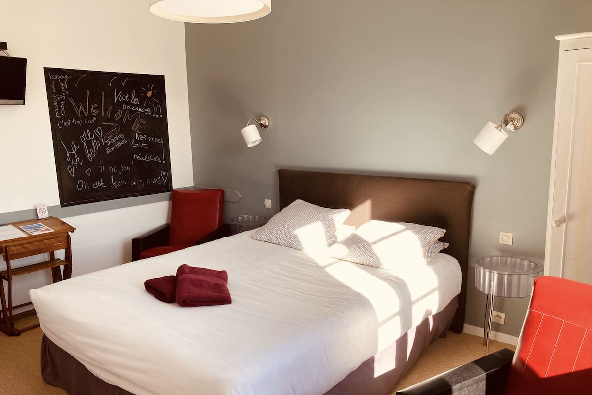 A neatly made bed with white linens and a red towel, a red chair, a small wooden table with a magazine, and a blackboard with various messages.