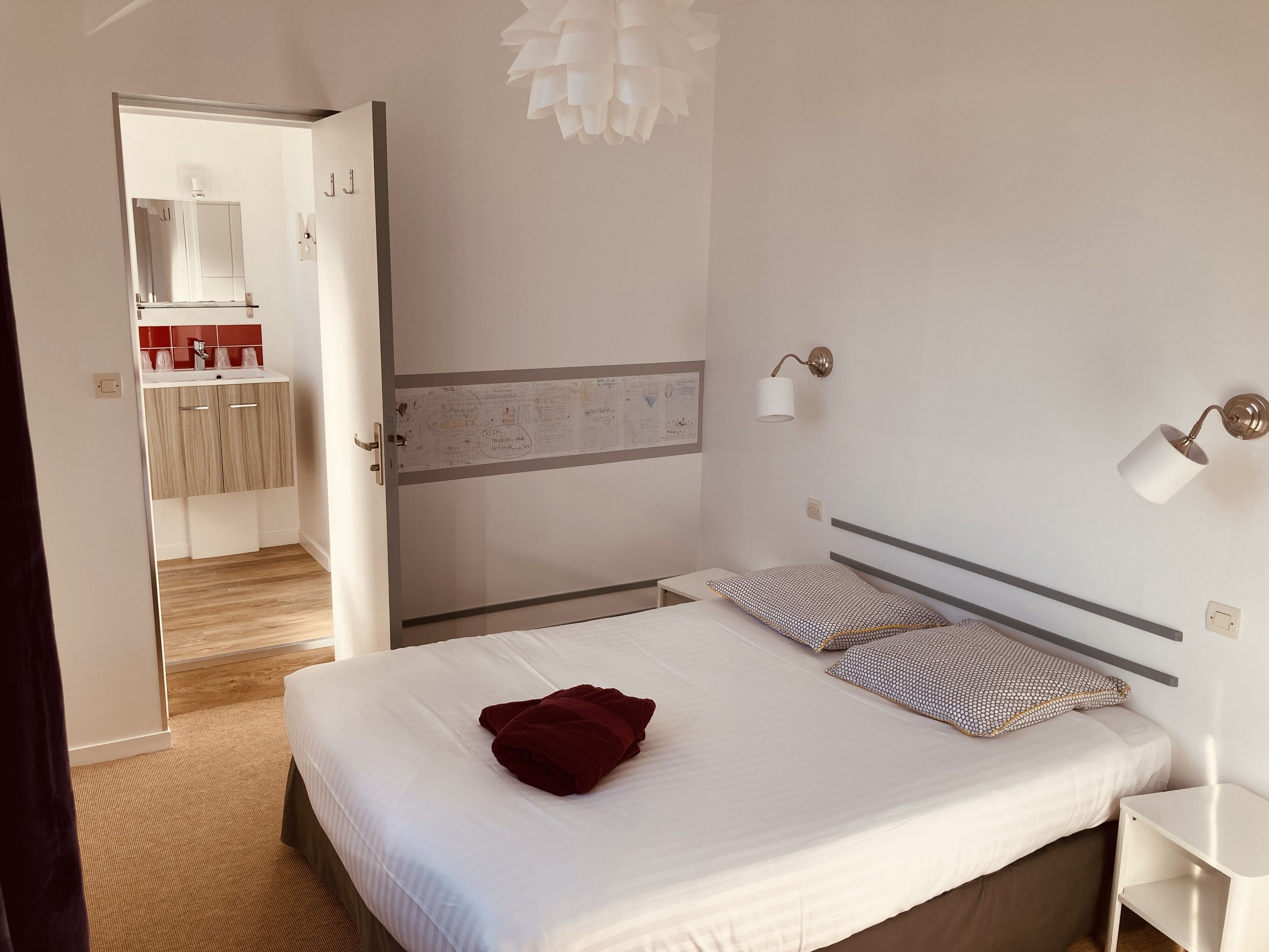 A hotel room with a bed, a bedside table, a nightstand, a wall-mounted lamp, and a mirror.