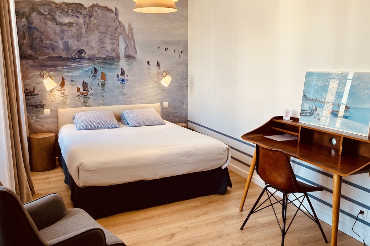 A hotel room with a large bed, a desk with a chair, a painting on the wall, and a hanging light fixture.