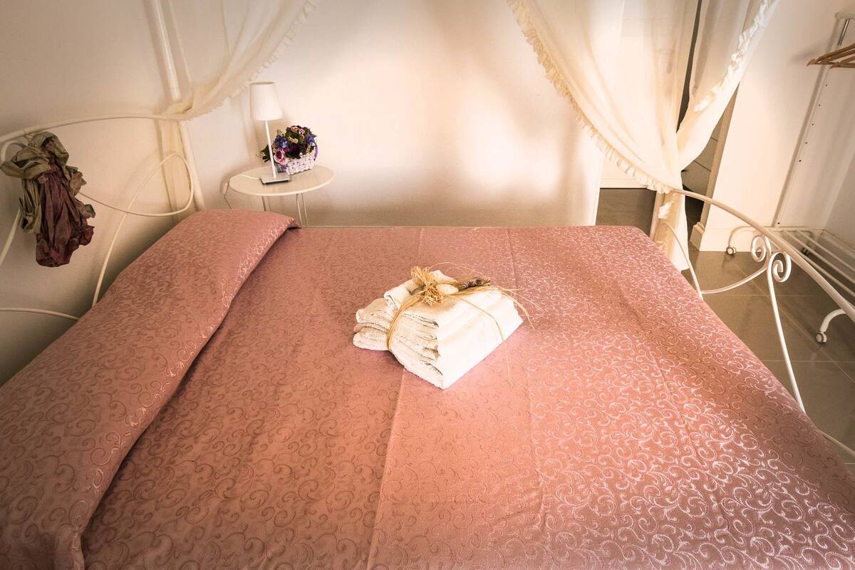 A neatly made bed with a pink patterned bedspread, a folded towel with a decorative bow, and a small table with a lamp and flowers.