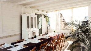 A dining area with a long table set for a meal, a white cabinet, and a large window with a view of a stone building. - B&B Il Borgo (Poggiardo)