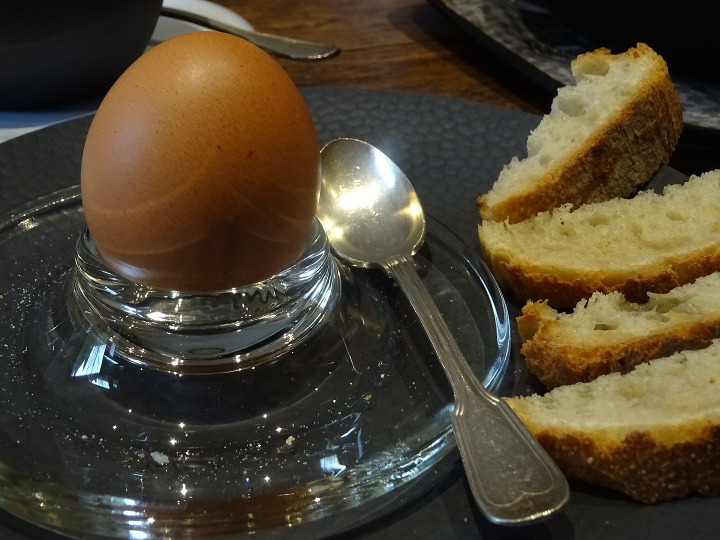A clear glass plate with a single egg on a stand, accompanied by a spoon and slices of bread.