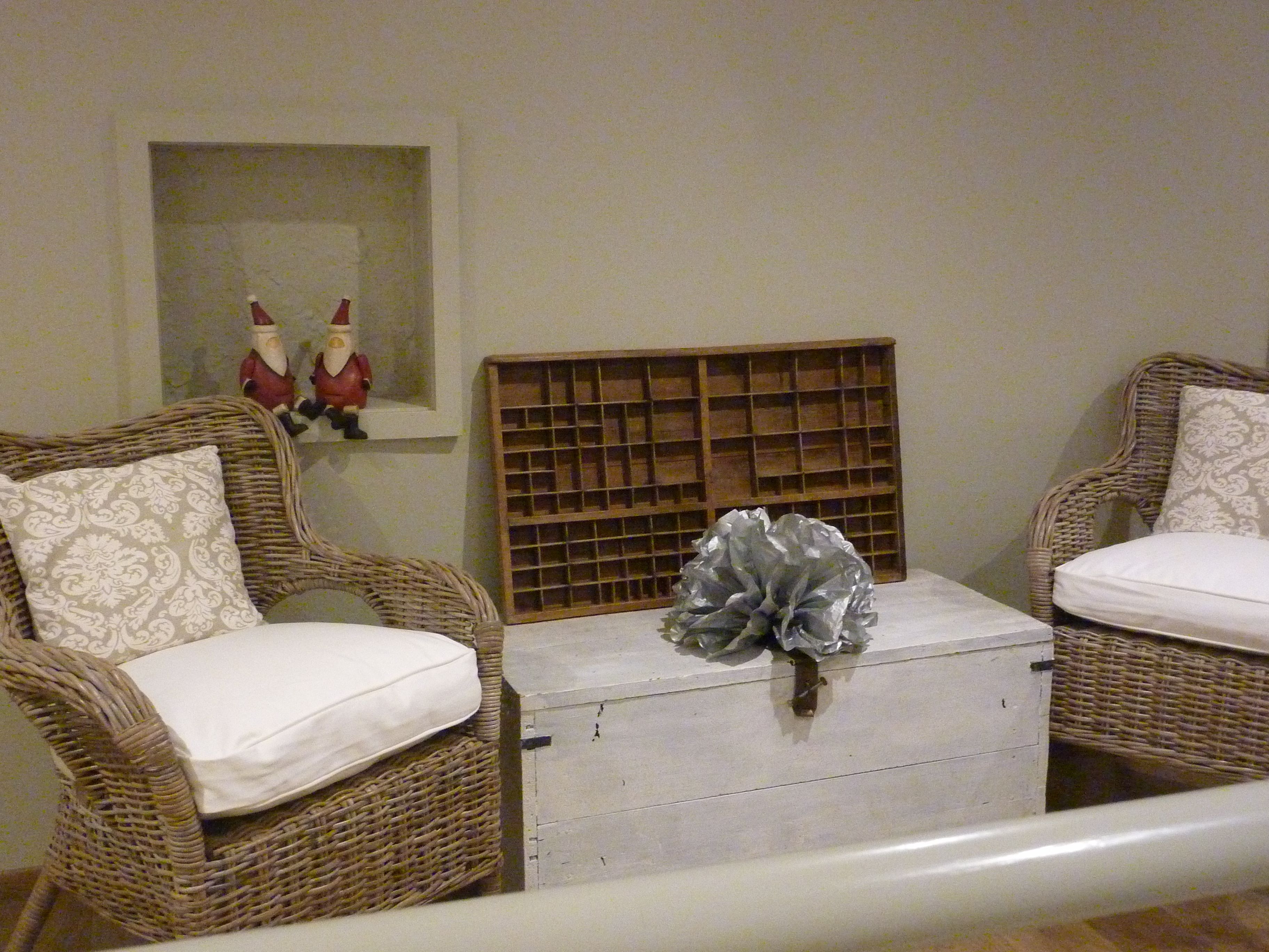 Une pièce comprenant deux fauteuils en osier avec des coussins blancs, une commode blanche ornée d’un nœud décoratif et une étagère en bois à claire-voie placée au-dessus.