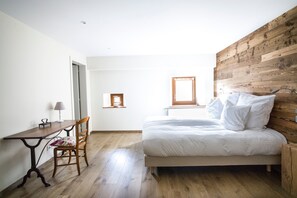 Un dormitorio con cabecera de madera, cama con ropa de cama blanca, un escritorio de madera con una silla y una ventana pequeña.