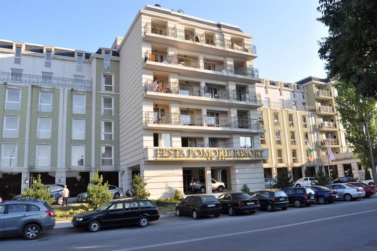 Un hotel de varios pisos con balcones, un letrero que dice 'FESTA POMORIE RESORT' y varios coches estacionados adelante.