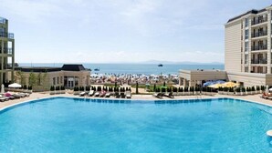 Una piscina con sillas de descanso y sombrillas, rodeada de edificios con balcones que dan a una playa y al mar.