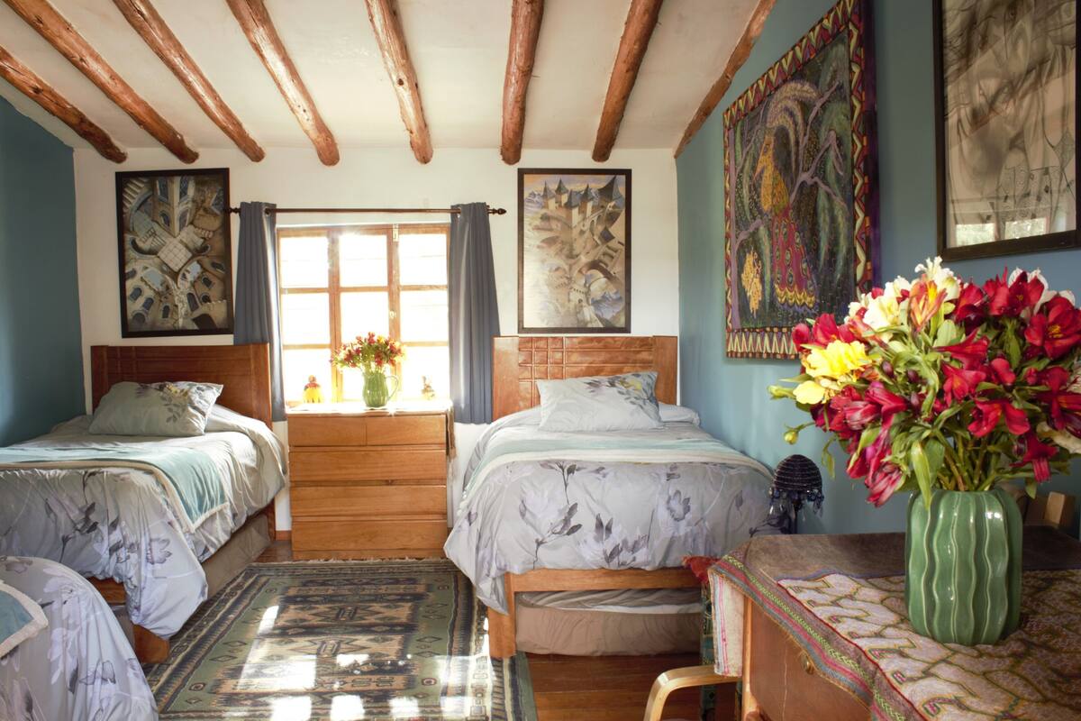 A bedroom with two beds, a wooden headboard, a window with curtains, and a vase with flowers on a table.