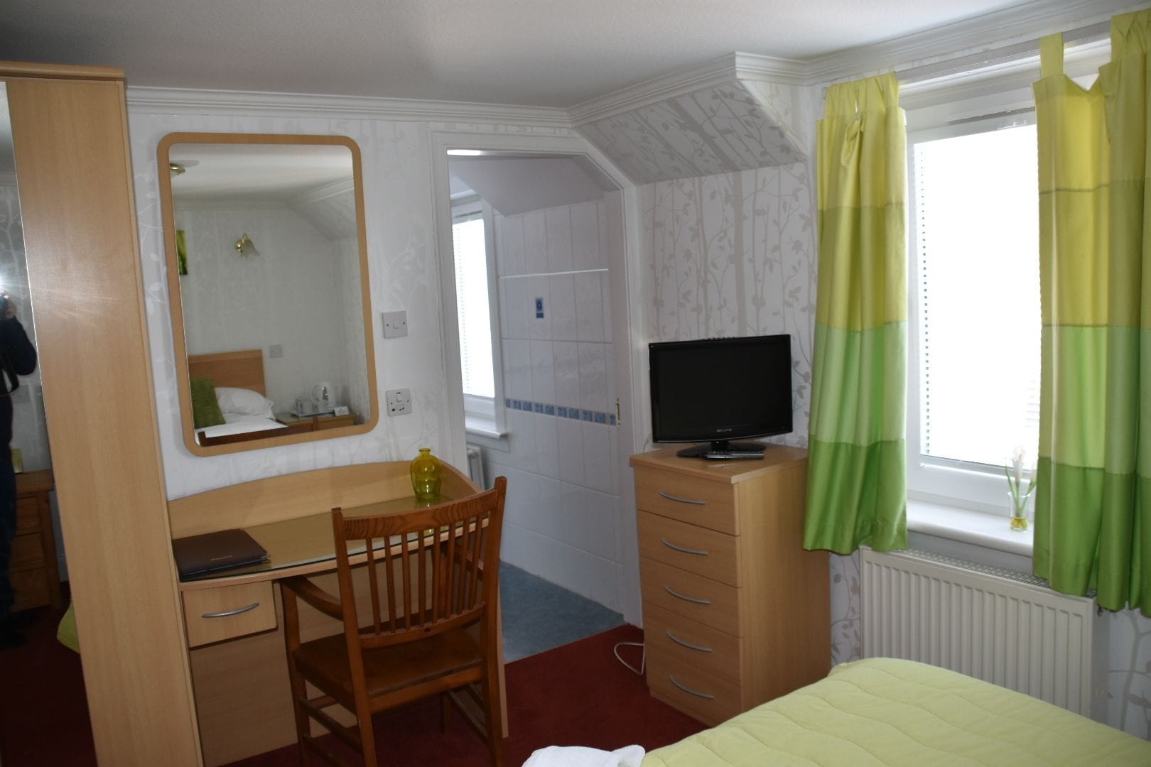 A hotel room with a bed, a desk with a chair, a television on a dresser, and a window with curtains.
